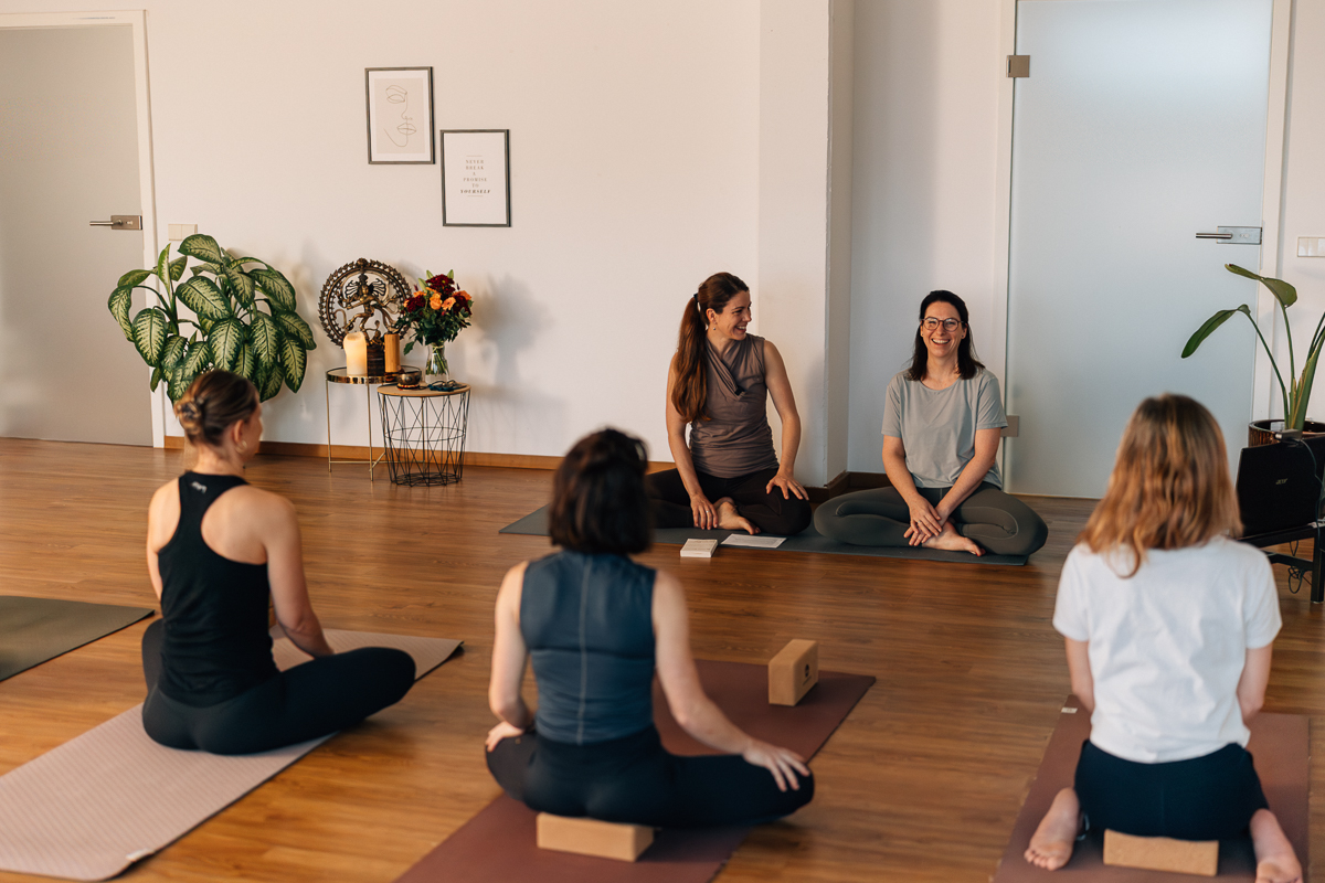 Stefanie Altendorf und Sabine Willmann, Yogalehrerinnen und Ausbilderinnen der Yoga Ausbildung München sitzen vor einer Gruppe und erzählen gut gelaunt von der nächsten Yoga Grundausbildung.